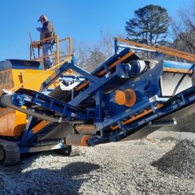 A large construction machine labeled "REBEL CRUSHER" is crushing rocks, with one man standing on top.
