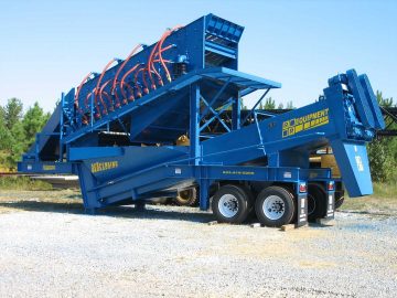 A blue industrial screening machine with multiple vibrational screens on a trailer, surrounded by trees.
