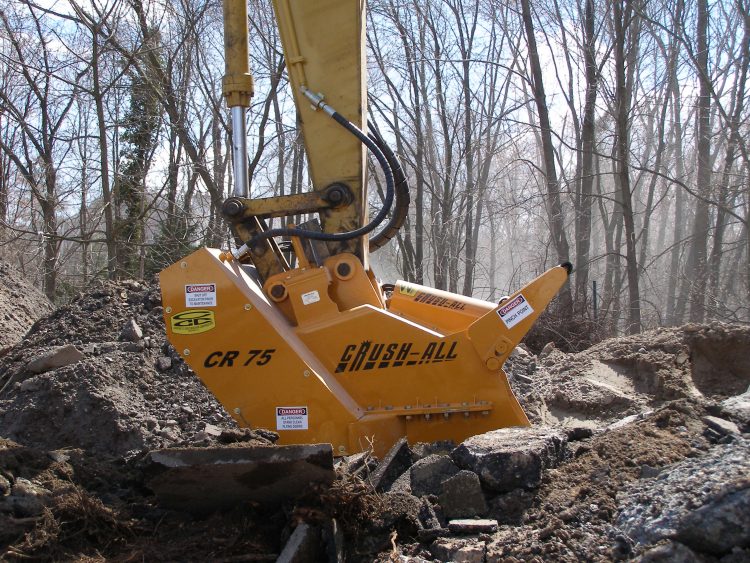 Excavator attachment labeled "CRUSH-ALL CR 75" actively breaks concrete debris amidst a wooded area.